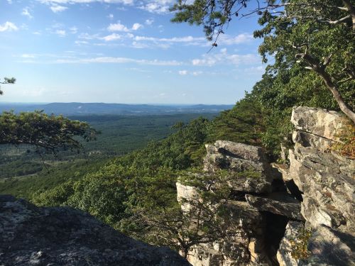 A scenic view of rolling hills and valleys from a rocky outcrop, with trees and a clear blue sky in the background.