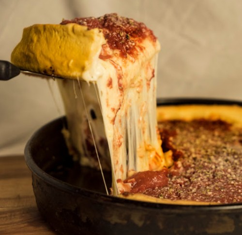 A slice of cheesy deep-dish pizza being lifted from a pan, with gooey cheese and tomato sauce stretching.
