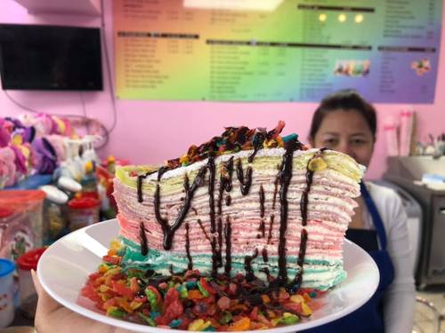 A colorful layered cake topped with chocolate sauce and colorful cereal, held in front of a shop counter.