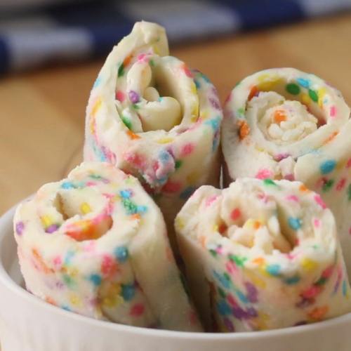 Colorful dessert rolls with sprinkles, arranged neatly in a white bowl on a wooden surface.