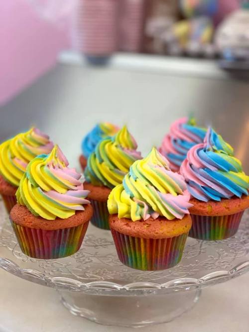 Colorful cupcakes with swirled pink, yellow, and blue frosting on a glass cake stand.