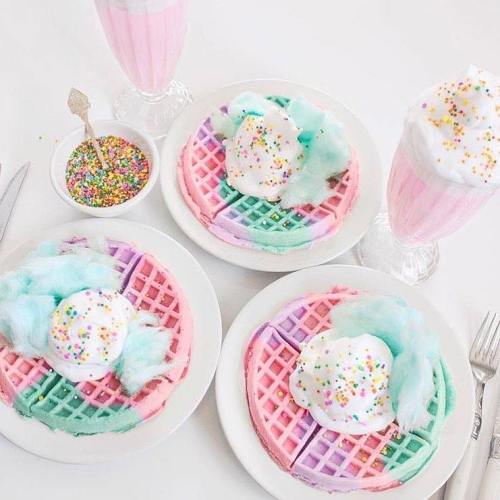 Three colorful waffles topped with cotton candy, whipped cream, and sprinkles, alongside pink milkshakes.