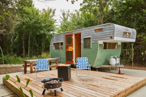 A vintage green camper trailer parked on a wooden deck, surrounded by trees, with outdoor seating and a fire pit.