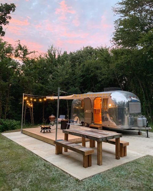 A shiny Airstream trailer with outdoor seating and string lights, set in a grassy area surrounded by trees at sunset.