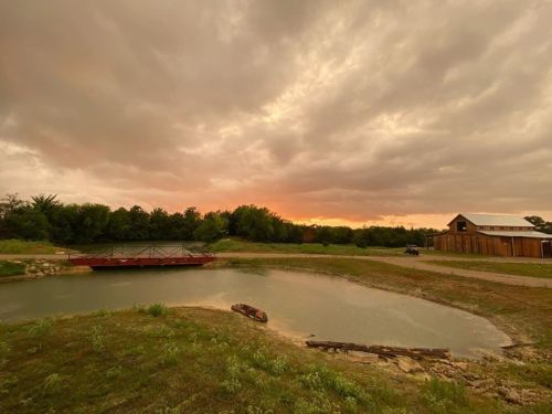 A serene landscape featuring a pond, a wooden structure, and a dramatic sunset behind cloudy skies.