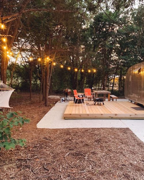 Cozy outdoor space with string lights, wooden deck, two chairs, and a hammock surrounded by trees at sunset.