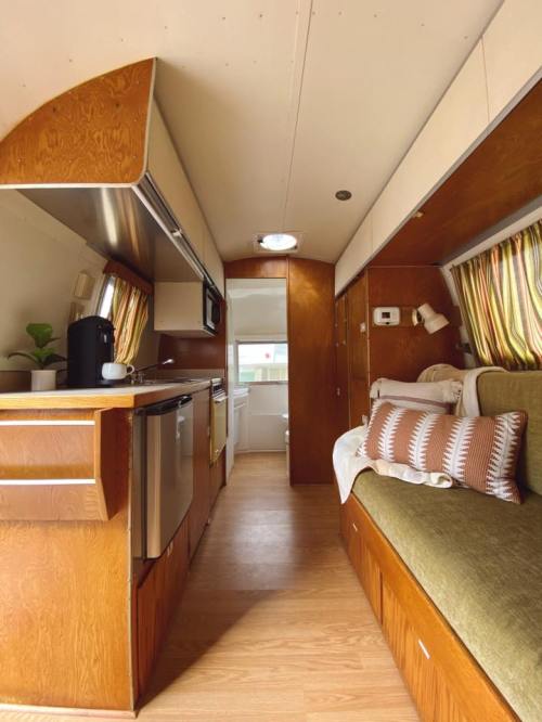 Cozy interior of a vintage camper, featuring a kitchen area, seating, and warm wood accents. Natural light filters in.