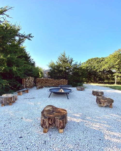 A fire pit surrounded by log seating in a gravel area, with trees and stacked firewood in the background.