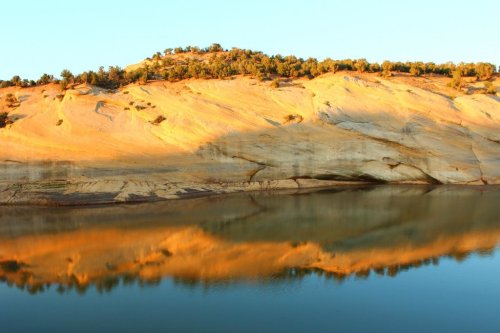 Golden-hued cliffs reflect in calm water under a clear blue sky, surrounded by lush greenery.