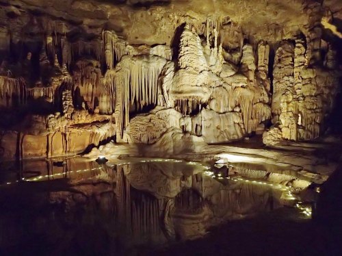 A dimly lit cave with stalactites and stalagmites reflected in a calm pool of water, illuminated by soft lights.