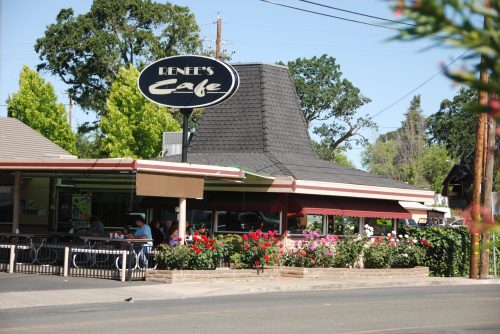 A cozy café with a sign reading "Renee's Café," surrounded by flowers and trees on a sunny day.