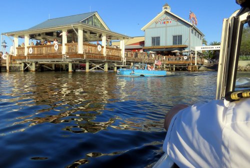 A view of a waterfront restaurant with a boat passing by on a sunny day. Reflections shimmer on the water's surface.