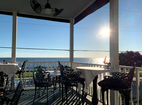 A scenic view from a porch with tables overlooking a calm sea and a bright sun in the sky.