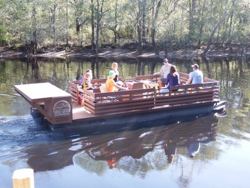 A group of people enjoying a boat ride on a calm river, surrounded by trees and reflecting water.
