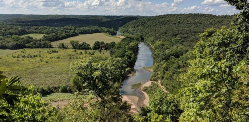 A scenic view of a winding river surrounded by lush green hills and fields under a partly cloudy sky.