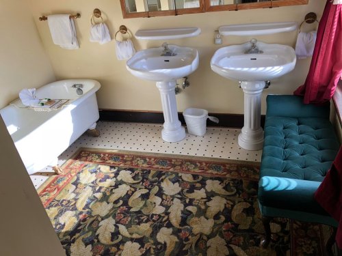 A bright bathroom featuring two sinks, a bathtub, a small couch, and a patterned rug.