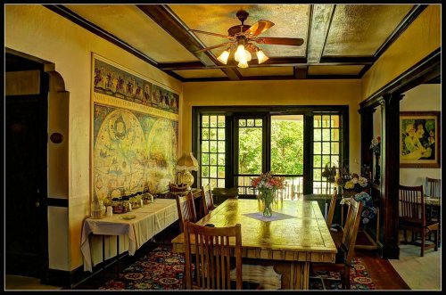 A cozy dining room with a large wooden table, decorative wall mural, and windows overlooking greenery.