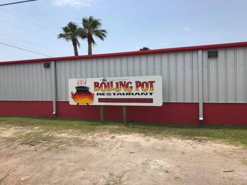 Sign for "The Boiling Pot Restaurant" with a graphic of a pot and flames, set against a metal building and palm trees.