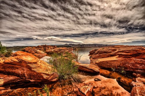 A scenic view of red rock formations by a calm lake under a dramatic, cloudy sky.