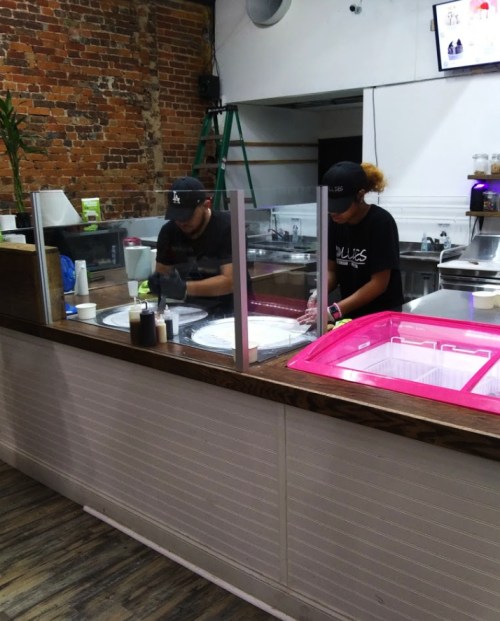Two workers prepare food behind a counter in a restaurant with a brick wall and a TV in the background.