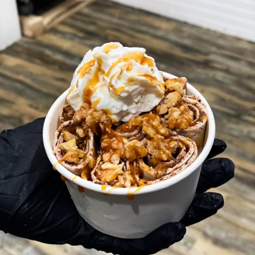 A hand holds a cup of rolled ice cream topped with whipped cream, nuts, and caramel drizzle.