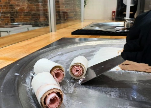 A chef's hand uses a spatula to roll ice cream into cylindrical shapes on a cold surface.