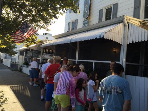 A line of people waiting outside a restaurant with an American flag in the foreground and a sunny atmosphere.