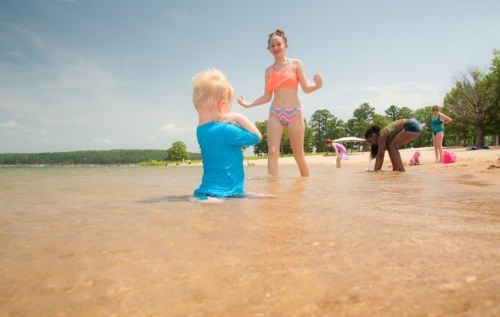 A child in a blue shirt plays in shallow water while a girl in a bikini stands nearby at a sunny beach.