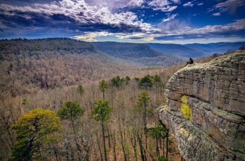 A person sits on a rocky ledge overlooking a vast forested valley under a dramatic sky.