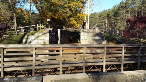 A wooden bridge leads to stone structures surrounded by trees with autumn foliage in a scenic landscape.
