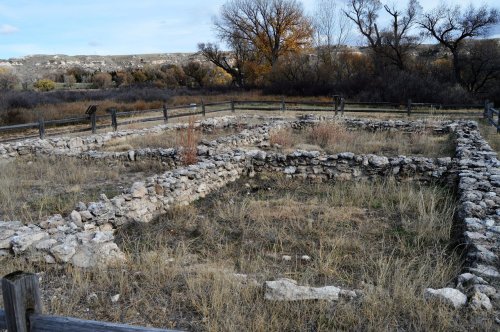 Ruins of a stone structure surrounded by grass and trees, with a wooden fence enclosing the area.
