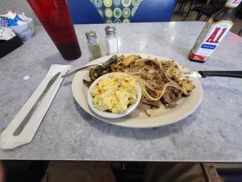 A plate of grilled meat with onions, a side of macaroni salad, and condiments on a table with a drink.