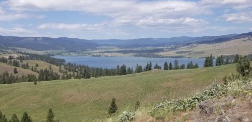 A scenic view of a lake surrounded by rolling hills and trees under a partly cloudy sky.