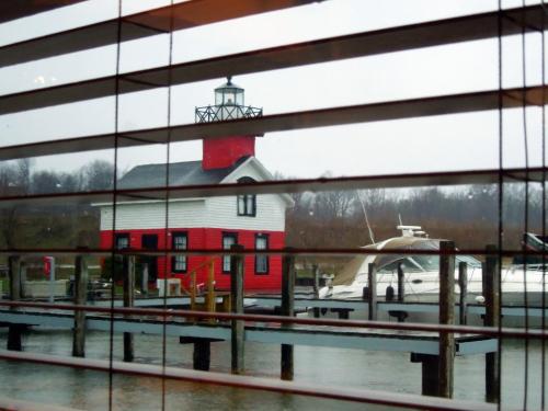 View of a red and white lighthouse by the water, seen through slatted blinds, with boats docked nearby.