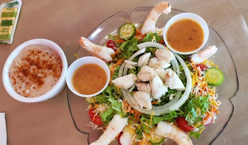 A colorful salad with shrimp, crab meat, and two dipping sauces, served on a clear plate with a side of seasoning.