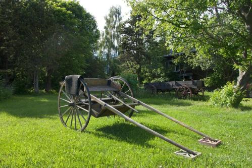 A wooden cart with long handles sits on green grass, surrounded by trees and another cart in the background.