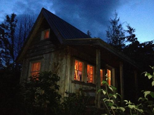 A cozy wooden cabin illuminated from within, surrounded by trees under a darkening sky.