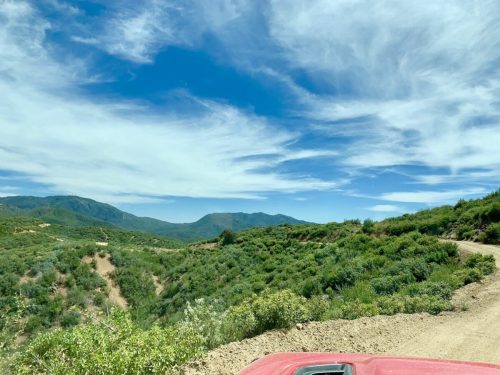 A scenic view of green hills and mountains under a blue sky with wispy clouds, seen from a dirt road.