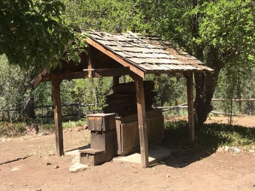 A rustic wooden shelter with a slanted roof, surrounded by trees and a dirt area, holding stacked materials inside.