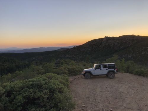 A white Jeep parked on a dirt path overlooking a mountainous landscape at sunset.
