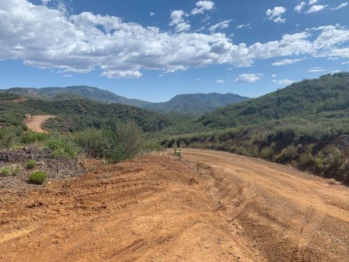 A winding dirt road through green hills under a blue sky with scattered clouds. Mountains are visible in the distance.