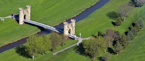 Aerial view of a stone bridge with towers over a river, surrounded by lush green grass and trees.