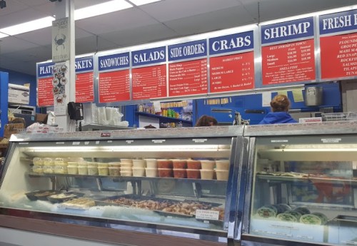A seafood market display case with various seafood items and a menu board featuring platters, sandwiches, and crabs.