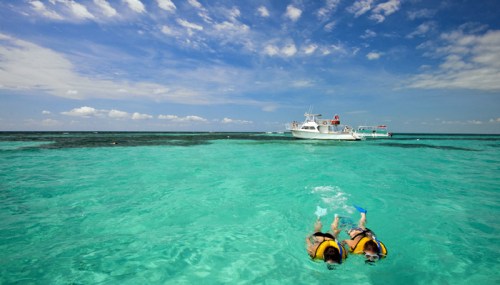 Two people in life jackets snorkeling in clear turquoise water, with a boat in the background under a blue sky.