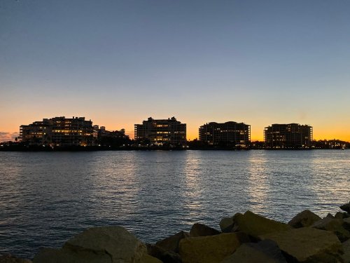 Silhouetted buildings along a waterfront at sunset, with calm water reflecting the evening sky.