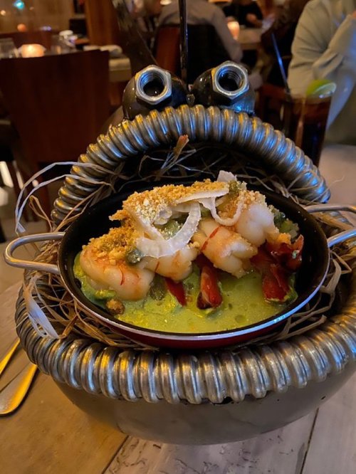 A whimsical seafood dish served in a decorative crab-shaped bowl, featuring shrimp and vibrant green sauce.