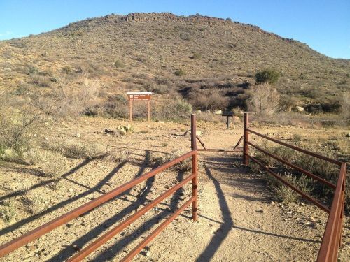 A dirt path leads to a hill, framed by metal fencing and signs, under a clear blue sky. Sparse vegetation surrounds the area.