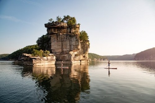 Two paddleboarders glide on calm water near a large rock formation surrounded by trees and mountains.