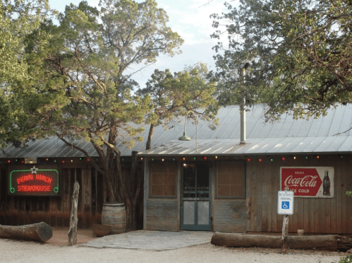 A rustic steakhouse with wooden exterior, Coca-Cola signs, and festive lights, surrounded by trees.
