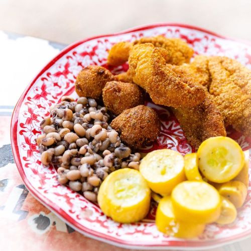 A vibrant plate featuring fried catfish, black-eyed peas, corn fritters, and sliced yellow squash on a decorative red plate.